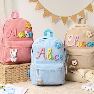May include: Three corduroy backpacks in pink, light blue, and beige. Each backpack features a name embroidered in colourful letters: Moira, Alice, and Elisa. The backpacks have a front pocket and a top handle. Decorative accents include a flower and a small stuffed animal.