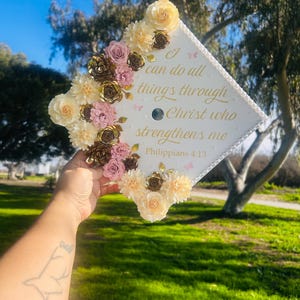 May include: A white graduation cap decorated with cream, pink, and gold paper flowers. The cap features the text "I can do all things through Christ who strengthens me" in gold lettering, with small butterfly accents.