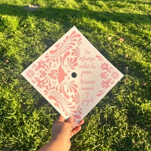 May include: A graduation cap with a white base and a pink floral design. The cap has the text "Su esfuerzo valió la pena. Gracias mamá y papá" in pink. The cap is held over a green grassy background.