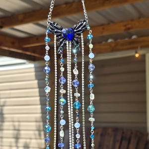 May include: A decorative hanging ornament featuring a dark metal butterfly design with a large blue heart-shaped gem. Strands of blue, clear, and iridescent beads and teardrop pendants hang below, interspersed with silver rhinestone chains.