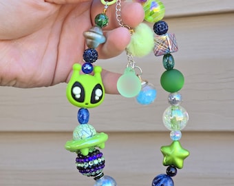 May include: A green alien charm with a spaceship charm, green, blue, and iridescent beads, and a green star charm on a beaded bracelet.