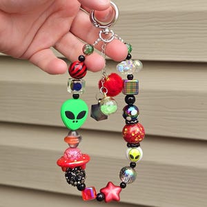May include: A colorful beaded keychain with a green alien, red UFO, and red star charm. The keychain is attached to a silver ring and chain.