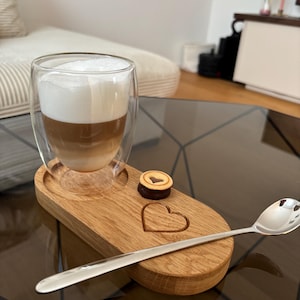 Könnte beinhalten: Ein doppelwandiges Glas mit geschichtetem Kaffeegetränk und einer dicken Schaumschicht, das auf einem Holztablett steht. Das Tablett hat eine herzförmige Vertiefung, einen kleinen Keks und einen silbernen Löffel.