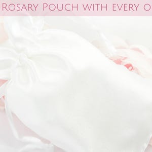 May include: A white satin rosary pouch with a drawstring closure, set against a backdrop of soft pink and white flowers. The text "FREE ROSARY POUCH WITH EVERY ORDER" is displayed at the top.