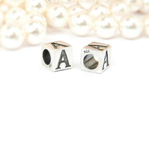 May include: Two silver cube charms, each featuring the letter "A" in a bold, black font. The charms are marked with "925" and have a hole for stringing. A blurred strand of white pearls is in the background.