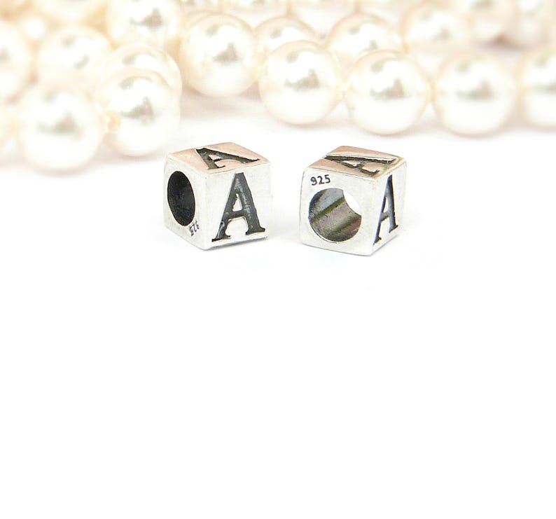 May include: Two silver cube charms, each featuring the letter "A" in a bold, black font. The charms are marked with "925" and have a hole for stringing. A blurred strand of white pearls is in the background.