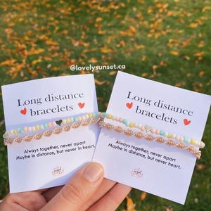 Op de afbeelding: Twee kaarten met de tekst "Long distance bracelets" en de zin "Altijd samen, nooit uit elkaar. Misschien op afstand, maar nooit in het hart." De kaarten worden in een hand gehouden en tonen twee armbanden met kleurrijke kralen.