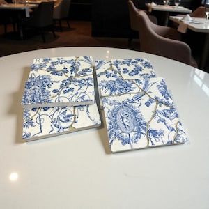 Könnte beinhalten: Vier quadratische Keramikuntersetzer mit blau-weißem Blumendesign und goldfarbenen Rissen im Kintsugi-Stil. Die Untersetzer sind auf einer weißen Oberfläche angeordnet und zeigen eine klassische Ästhetik.