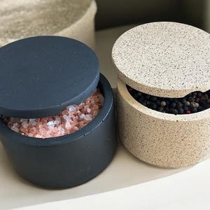 May include: Two round, lidded salt and pepper cellars. One is dark grey and filled with pink salt crystals. The other is speckled beige and contains peppercorns. A third, similar container is in the background.