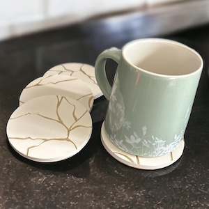 May include: Four white ceramic coasters with a gold crackle design and a green ceramic mug with a white floral design on a black surface.