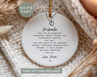 May include: A round white ceramic ornament featuring a black line drawing of a heart pendant on a chain at the top. The text reads: Mikaela, You are my greatest blessing. Never forget how incredibly special you are. You are born to do amazing things. Continue chasing your dreams. I will always be here cheering you on. Love, Mom, with a small heart icon below.