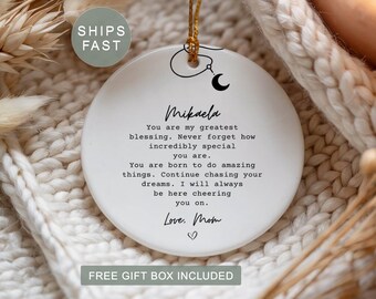 May include: A round white ceramic ornament featuring a black printed message that reads: Mikaela, You are my greatest blessing. Never forget how incredibly special you are. You are born to do amazing things. Continue chasing your dreams. I will always be here cheering you on. Love, Mom. It includes a small black crescent moon graphic and a gold hanging string.