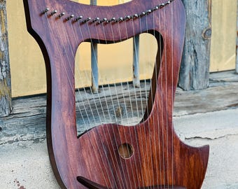 May include: A wooden lyre with a dark brown stain. The lyre has 15 strings and a sound hole in the center. The lyre is resting on a concrete surface.