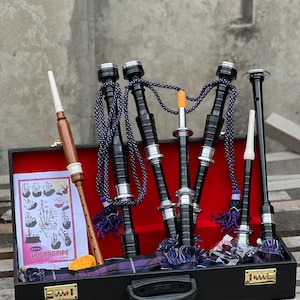 Puede incluir: Un conjunto de gaita negra con interior rojo y tela de tartán morado y negro. El conjunto de gaita incluye un chanter, un fuelle y tres drones. El conjunto de gaita está en una maleta de transporte negra con una cerradura dorada.