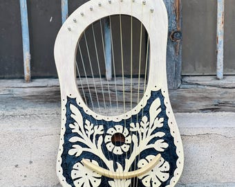 Op de afbeelding: Een houten lier met een lichtgekleurd frame en een donker, sierlijk bloemmotief. Het instrument heeft 16 snaren en rust op een verweerd oppervlak. Het ontwerp is geschilderd in zwart en wit.