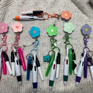 May include: A collection of colorful retractable pens attached to flower-shaped badge reels. The pens have various colored accents, and the badge reels come in pink, blue, green, and purple. The background is a textured, light gray fabric.