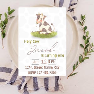 May include: A white and black cow with brown eyes and a pink nose is eating grass. The text "Holy Cow Jacob is turning one" is written in brown cursive font. The date "Jan 12 5 PM" and the address "1234, Street Name, City" are written in black font. The text "RSVP 123 456 7890" is written in brown font. This is a birthday invitation.