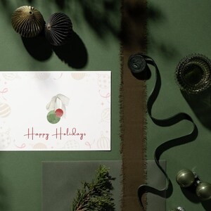 May include: A white card with a watercolor illustration of a green and red Christmas ornament with the text "Happy Holidays" in red cursive.