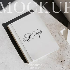 May include: A white book with the word "Mockup" in elegant script on the cover. The book is on a textured surface, with the word "Mockup" in large, faded letters above it. The Etsy shop name "svoemisto" is at the bottom.