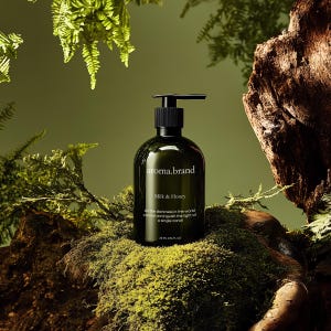 Puede incluir: Una botella de vidrio verde oscuro con una tapa de bomba negra y una etiqueta que dice "aroma.brand Milk & Honey". La botella está sentada sobre un lecho de musgo verde. El texto de la etiqueta dice "Toda la oscuridad del mundo no puede extinguir la luz de una sola vela 25 ml 0.84 fl oz".