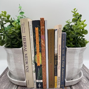 May include: A set of two speckled white ceramic planters with attached saucers, each containing a green faux plant. Between the planters is a row of nine books with various titles including Herbal Medicine, Gathering Moss, and The Four Agreements.