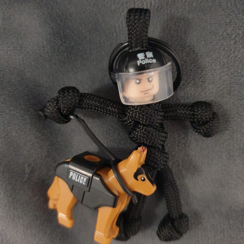 Puede incluir: Un llavero de paracord negro con un polic&iacute;a de estilo Lego con una visera transparente y un perro polic&iacute;a de estilo Lego. El perro es de color marr&oacute;n y negro, con un chaleco que dice "POLICE". El casco del oficial tiene la palabra "Police".