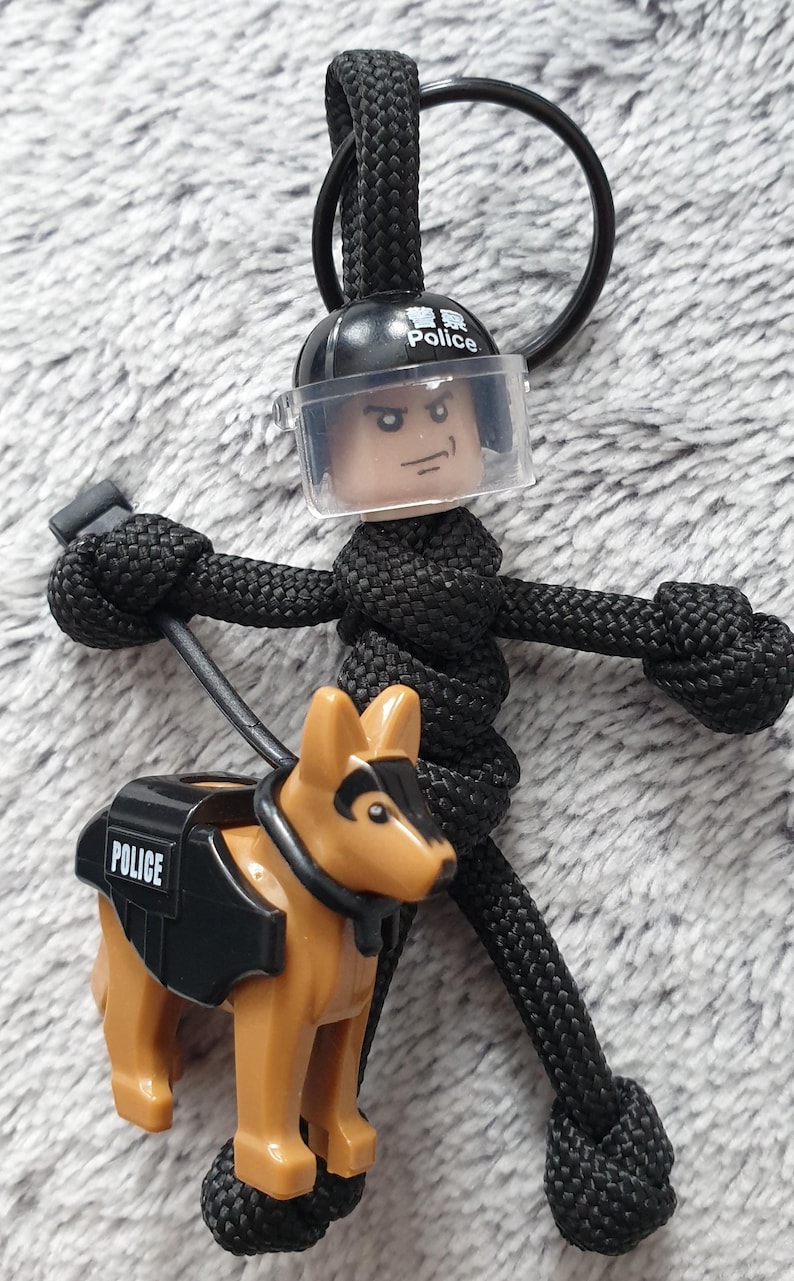 Puede incluir: Llavero de paracord negro con una figura de polic&iacute;a con visera transparente y un perro polic&iacute;a pastor alem&aacute;n. El perro tiene un chaleco negro con "POLICE" impreso. El casco del oficial tiene "Police" impreso.