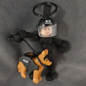 Puede incluir: Llavero de paracord negro con un polic&iacute;a Lego con una visera transparente y un perro polic&iacute;a Lego. El casco del oficial dice "Police" en ingl&eacute;s y chino. El perro lleva un chaleco negro que dice "POLICE".