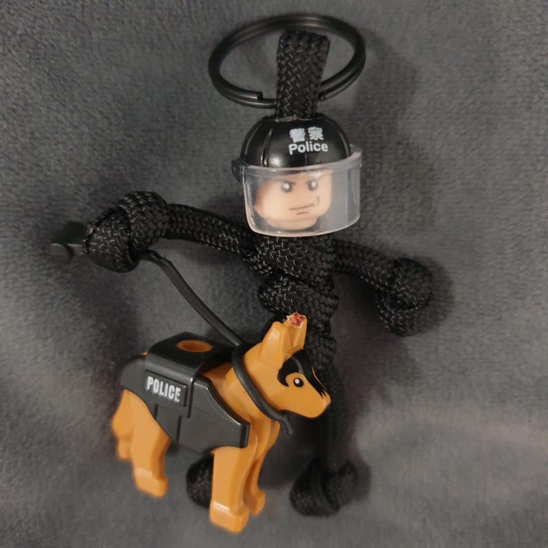 Puede incluir: Llavero de paracord negro con un polic&iacute;a Lego con una visera transparente y un perro polic&iacute;a Lego. El casco del oficial dice "Police" en ingl&eacute;s y chino. El perro lleva un chaleco negro que dice "POLICE".