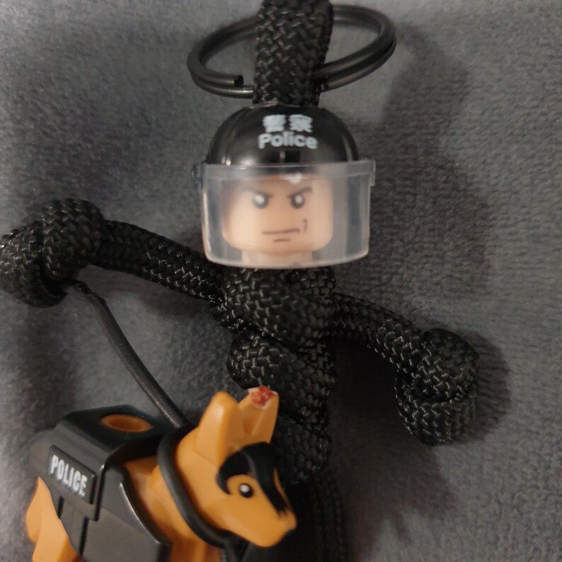 Puede incluir: Un llavero de paracord negro con un polic&iacute;a de Lego con una visera transparente y un perro polic&iacute;a de Lego. El casco del oficial tiene la palabra "Police" en ingl&eacute;s y caracteres chinos. El llavero tiene un anillo de metal.