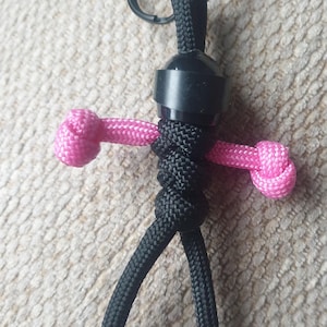 Może przedstawiać: Czarno-różowy brelok z paracordu z czarnym metalowym kółkiem. Brelok ma kształt ludzika z ramionami i nogami.