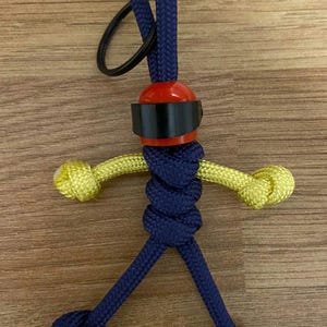 May include: A blue and yellow paracord keychain with a red and black helmet charm. The keychain is made of braided cord and has a loop at the top for attaching to keys.