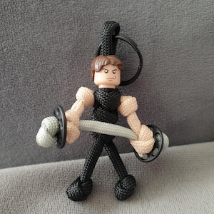 Könnte beinhalten: Eine schwarze und braune Paracord-Figur mit einem braunen Kopf und einer silbernen Langhantel. Die Figur hält die Langhantel mit beiden Händen und hat einen schwarzen Ring, der an der Oberseite des Kopfes befestigt ist.