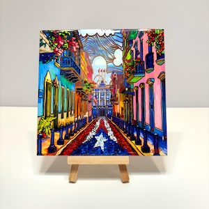 Puede incluir: Una pintura colorida de una escena callejera en el Viejo San Juan, Puerto Rico. La calle está bordeada de edificios de colores brillantes y una calle adoquinada con el diseño de la bandera puertorriqueña. La pintura está en un pequeño caballete.