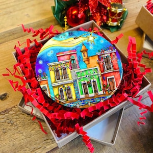 Puede incluir: Adorno navideño redondo con una colorida ilustración de edificios con balcones y luces festivas. El adorno está en una caja de regalo con papel rojo, lo que sugiere un tema navideño. Los edificios están pintados con colores vibrantes.
