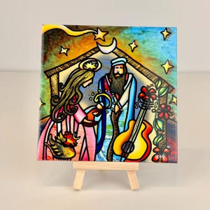 May include: A square ceramic tile with a stained-glass effect, depicting a nativity scene. The image shows Mary, Joseph, a guitar, and a rooster, set against a starry sky and a crescent moon. The tile is displayed on a small wooden easel.