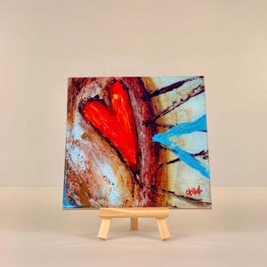 May include: A square canvas painting on a small wooden easel. The artwork features a large red heart in the center, with blue and brown brushstrokes. The painting has a textured appearance and is signed in the bottom right corner.