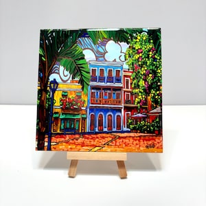 May include: A colorful painting of a street scene in a tropical city. The buildings are painted in bright colors, including blue, yellow, red, and green. There are palm trees and lush foliage in the foreground. The painting is on a small easel.