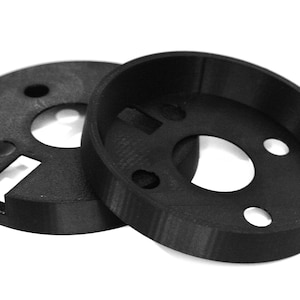 Puede incluir: Dos anillos de plástico negro con agujeros de diferentes tamaños en el centro. Los anillos están diseñados para apilarse uno encima del otro.