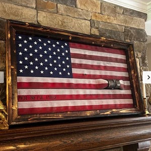 May include: A rectangular wall art piece featuring a United States flag design constructed from horizontal red and white fire hoses with a blue canton of white stars. The flag is encased in a dark, scorched-finish wooden frame with text reading PER NFPA 1962.