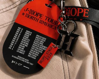 May include: A red and black tour pass for J-Hope Tour in North America with a list of dates and venues. Attached is a black metal H-shaped charm and a small red rectangular tag labeled Hope Stage 2025, all connected by a silver metal keyring.
