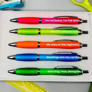 May include: A collection of five brightly coloured pens, each with a different witty phrase, presented in a white box. The pens are in shades of pink, green, orange, and blue. Phrases include "World's okayest boss" and "Warning: may delegate."