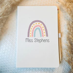 May include: A white notebook with a rainbow design and the text "Miss Stephens" on the front.