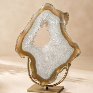 May include: A decorative geode slice featuring a translucent white crystalline center surrounded by a smooth, tan-colored agate border. The geode is mounted vertically on a brushed gold-toned metal stand with a square base and a U-shaped support.