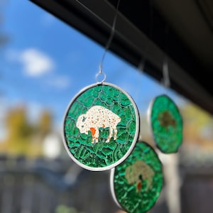 May include: Three round, green stained glass ornaments with silver frames. The ornaments feature a white buffalo, a tree, and a bird.