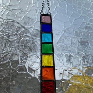 May include: A stained glass suncatcher with a rainbow of colors, including pink, blue, turquoise, green, yellow, orange, and red. The suncatcher is hanging from a chain.