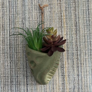 May include: A green ceramic planter shaped like a leaf with a hanging loop. The planter is filled with a green succulent and a purple succulent.