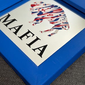 May include: A blue framed mirror with a white background. The mirror features a red, white, and blue abstract buffalo silhouette and the word "MAFIA" in black lettering.