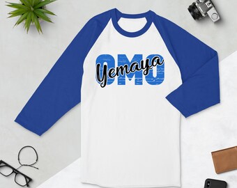 Puede incluir: Camiseta de béisbol raglán blanca y azul con las palabras "OMO" en azul y "Yemaya" en escritura negra. La camiseta tiene mangas azules y un cuerpo blanco. Una cámara, gafas y una pequeña planta están en el fondo.