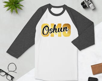 Puede incluir: Una camiseta de béisbol blanca y gris con un gráfico dorado y negro que dice "Oshun".